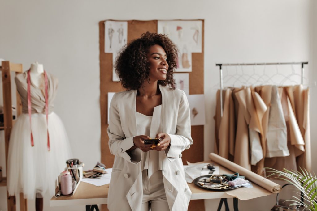 attractive dark skinned curly brunette woman oversized jacket white pants smiles holds phone leans table office fashion designer 2